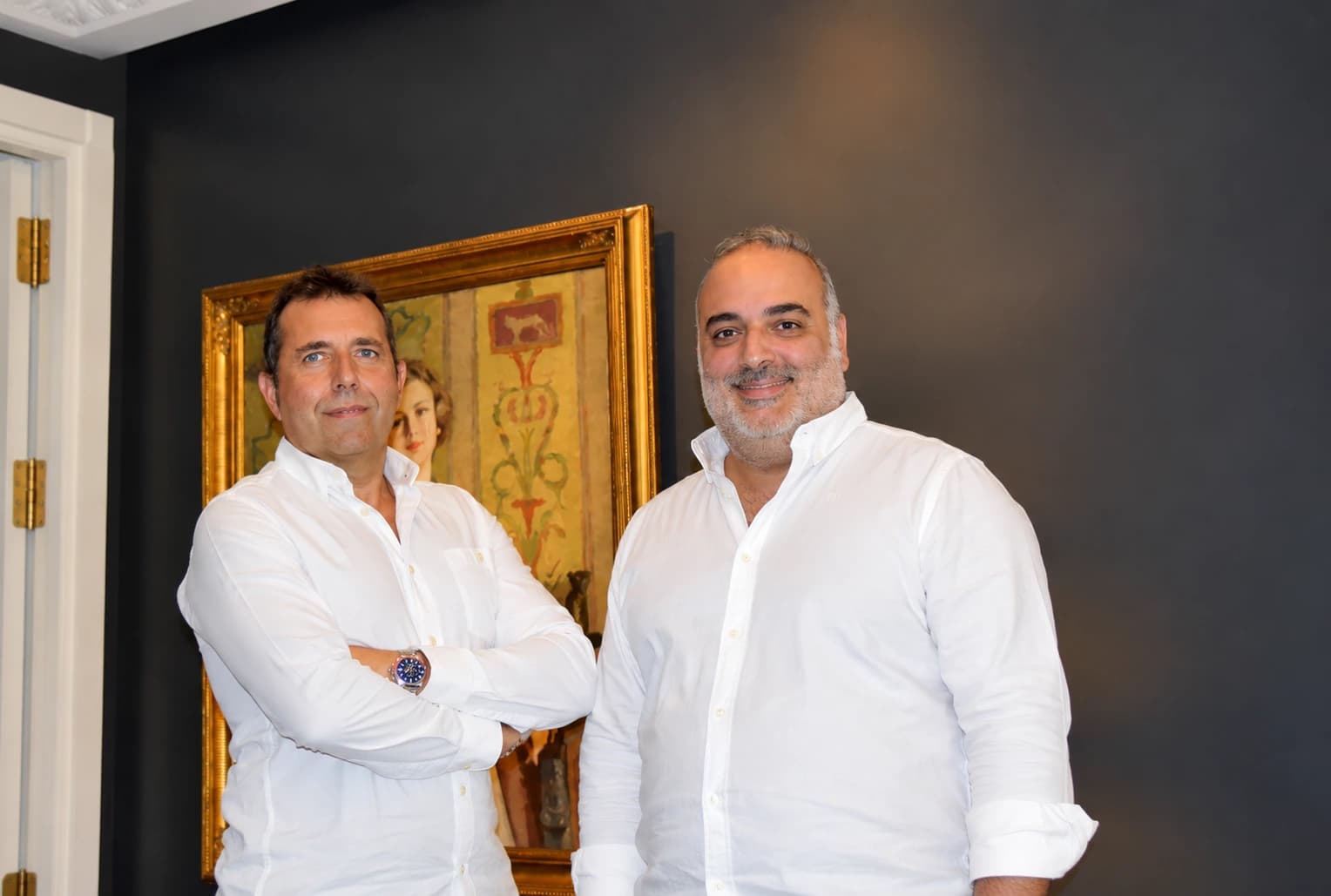Juan Luis y Rubén, socios de Clapham+Partners, con camisa blanca ante una pared oscura y un óleo con marco dorado